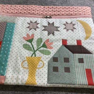 May include: A white zippered pouch with a patchwork design featuring a house, flowers in a vase, stars, and a crescent moon. The fabric is a mix of green, red, yellow, and brown.