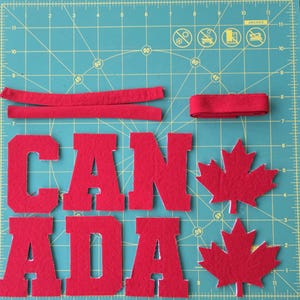 May include: Red felt letters spelling "CANADA" with two red maple leaf shapes and red felt strips on a green cutting mat. The letters are in a bold, block style.