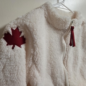 May include: A white fleece jacket with a high collar and a full zip. A burgundy maple leaf patch is sewn on the left shoulder. The zip pull has burgundy fabric accents.