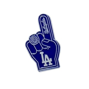 May include: A blue and white foam finger pin featuring the Los Angeles Dodgers logo and text. The design depicts a hand with an extended index finger, a circular ring detail with the LA emblem, and the words Los Angeles written vertically.