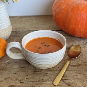 May include: A white ceramic mug with a handle filled with orange pumpkin soup. The soup is topped with fresh herbs. A whole orange pumpkin and a spoon are on the table next to the mug.