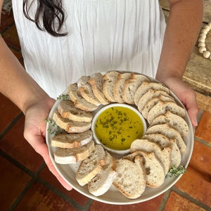 May include: A white ceramic plate with a small white bowl in the center filled with olive oil and herbs. The plate is surrounded by slices of crusty bread.