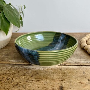 May include: A green and blue ceramic bowl with a striped design. The bowl is sitting on a wooden table.