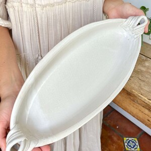 May include: White ceramic serving dish with two handles. The dish is oval shaped and has a slightly textured surface.