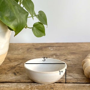 May include: A small white ceramic bowl with a 3-inch diameter and 1-inch height. The bowl is sitting on a wooden surface.