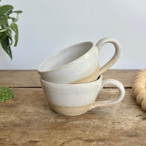 May include: Two handmade ceramic mugs stacked on top of each other. The mugs are off-white with a tan band at the base and a curved handle. The mugs are on a wooden surface. The mugs are part of a kitchenware set.