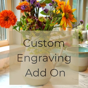 May include: A white ceramic pitcher with a brown rim, filled with a bouquet of colorful flowers. The text "Custom Engraving Add On" is superimposed on the image.