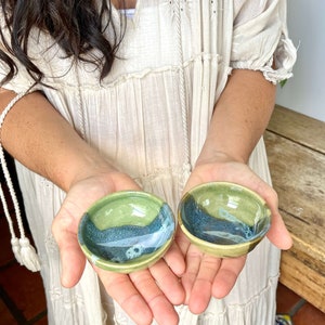 May include: Two small ceramic bowls with a green and blue glaze. The bowls are in the hands of a person.
