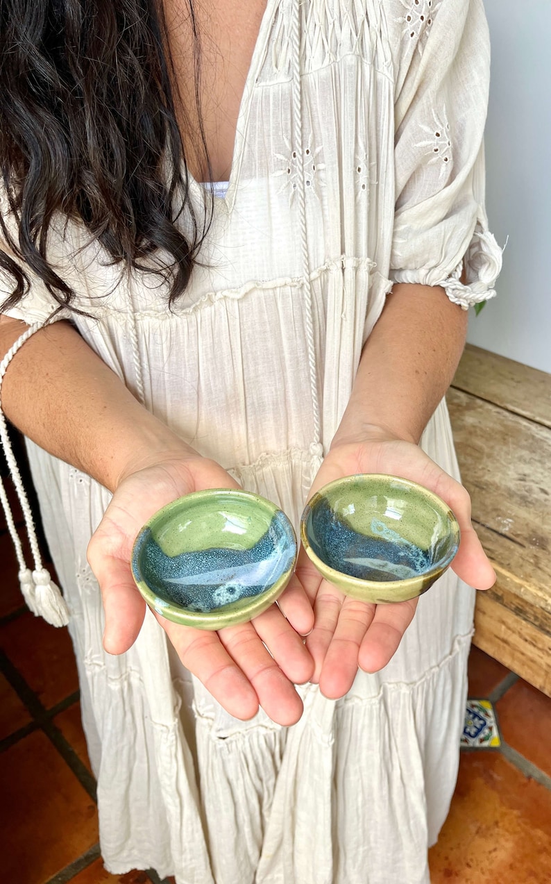 May include: Two small ceramic bowls with a green and blue glaze. The bowls are in the hands of a person.