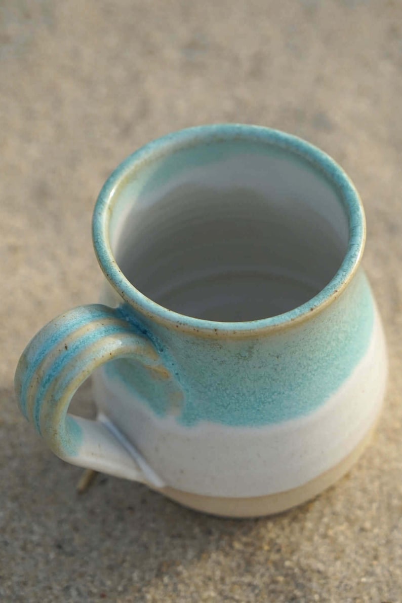 May include: A white ceramic mug with a light blue and beige glaze. The mug has a rounded body and a handle.