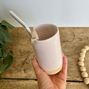 May include: A handmade, light pink ceramic cup with a rounded base and a spout. A clear glass straw is inserted into the cup. The cup is held in a hand against a wooden surface. The bottom of the cup is a light tan color.