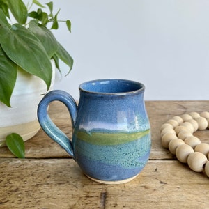 May include: A blue and green ceramic mug with a rounded body and a handle. The mug has a light blue glaze with a green stripe and a white stripe.