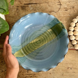 May include: A handmade ceramic serving bowl with a wavy rim. The bowl features a light blue base with a diagonal stripe of green and yellow concentric circles. The bowl is held by a hand, showcasing its size and design.