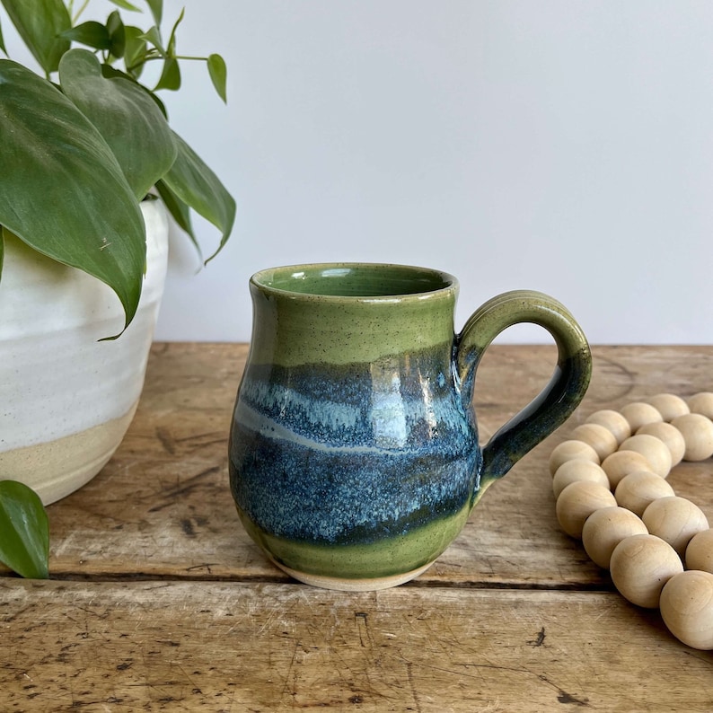 Op de afbeelding: Een groene en blauwe keramische mok met een rond lichaam en een handvat. De mok heeft een gespikkelde groene glazuur met een blauw en wit streepdessin.