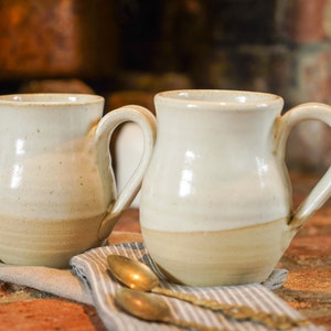 Op de afbeelding: Twee witte keramische mokken met bruine accenten en afgeronde bodems. De mokken hebben een gespikkeld glazuur en staan op een grijs-wit gestreepte doek.