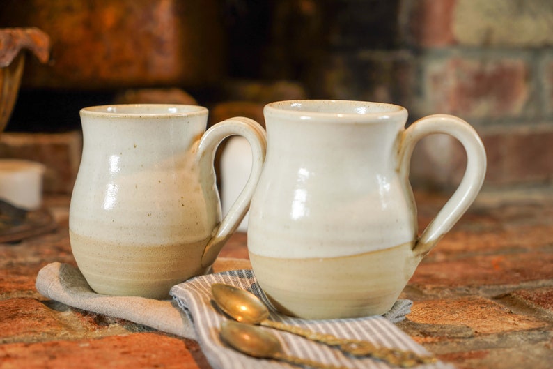 Op de afbeelding: Twee witte keramische mokken met bruine accenten en afgeronde bodems. De mokken hebben een gespikkeld glazuur en staan op een grijs-wit gestreepte doek.