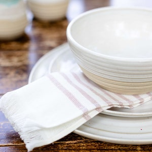 May include: A white ceramic bowl and plates are stacked on a wooden table. A white napkin with pink stripes and fringe is placed between the plates. The bowl has a textured, ridged design.