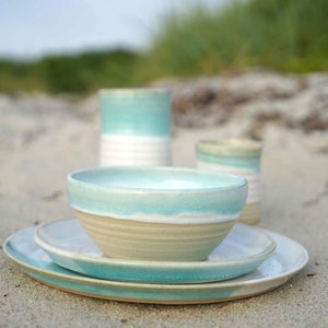 May include: A set of ceramic dishes in shades of blue and white, with a bowl, plate, and two cups. The dishes are arranged on a sandy surface.