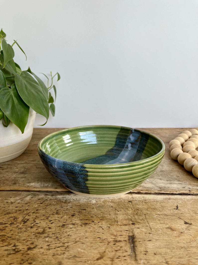May include: A green and blue ceramic bowl with a striped design. The bowl is sitting on a wooden table.