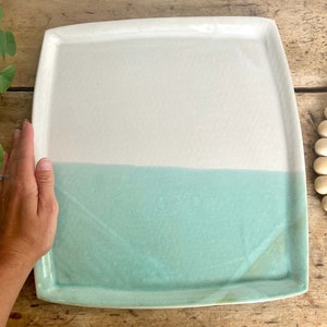 May include: A square ceramic serving platter with a white top and a light blue bottom. The platter has a slightly rounded edge.
