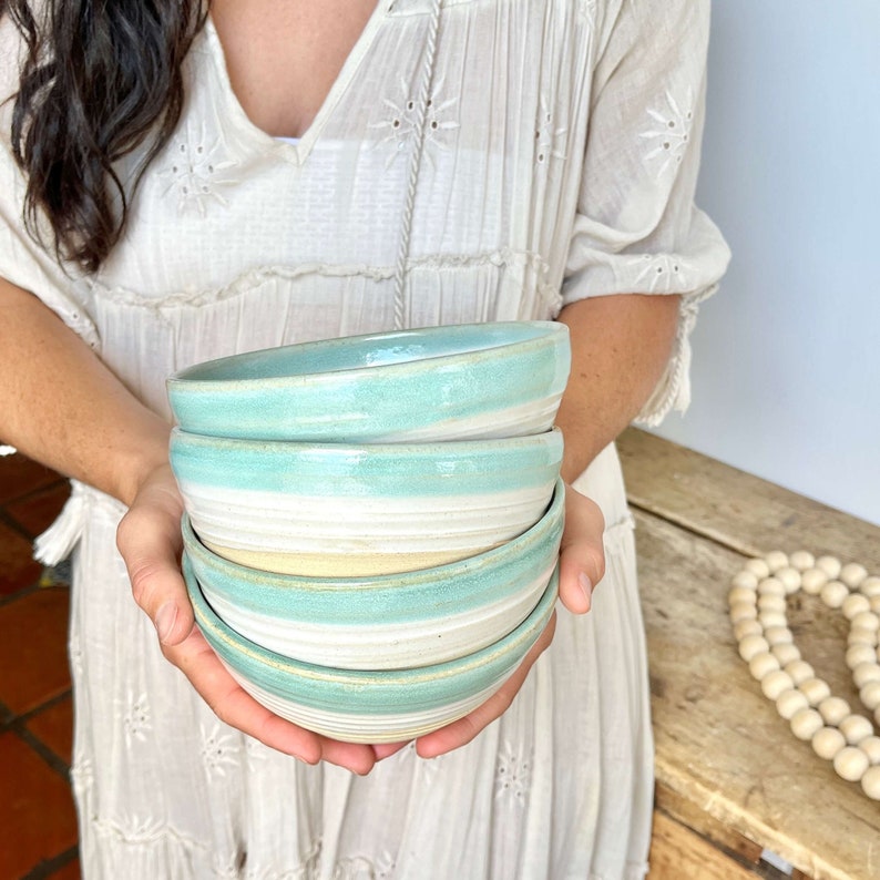 May include: A stack of four handmade ceramic bowls. The bowls have a light blue and white color scheme with a textured, glazed finish. The bowls are held in front of a person wearing a white dress.