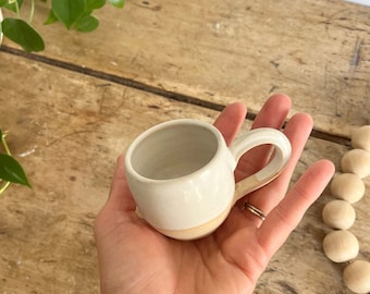 May include: A small, white ceramic mug with a rounded shape and a handle. The mug has a light brown band around the bottom.