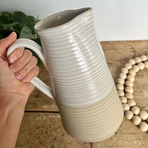 May include: A white ceramic pitcher with a textured, ribbed design. The pitcher has a wide mouth and a handle. The bottom half of the pitcher is a lighter shade of beige.