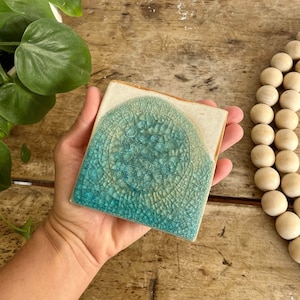 Peut inclure: Carreau de céramique carré avec un motif de glaçure craquelée turquoise et crème. La tuile est tenue dans une main, mettant en valeur la texture et les variations de couleur. La tuile a un aspect artistique unique.
