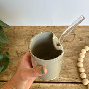 May include: A handmade ceramic mug in a muted green-gray color, featuring a unique curved design with a built-in straw holder. A clear glass straw is inserted into the mug. The mug is held by a person's hand. The mug sits on a wooden surface.