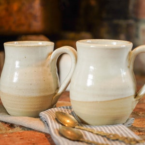 May include: Two white ceramic mugs with a brown glaze at the bottom. The mugs have a rounded body and a handle.