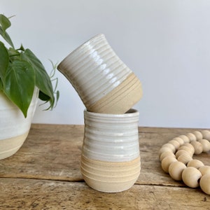 May include: Two white ceramic mugs with a beige band around the bottom. The mugs are stacked on top of each other.