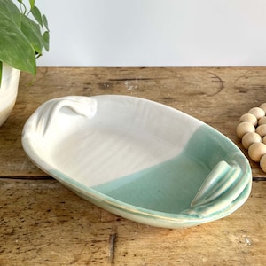 May include: A white and light green ceramic serving dish with two handles. The dish is oval shaped and has a glazed finish.