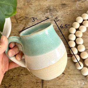 Op de afbeelding: Een lichtblauwe, witte en beige keramische mok met een handvat. De mok is 4,5 inch hoog en 3 inch breed.