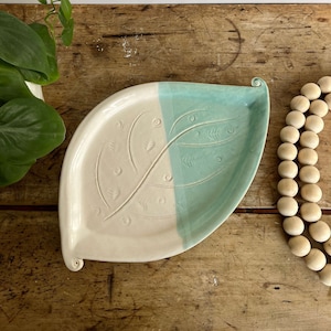 May include: A white ceramic leaf-shaped dish with a teal glaze on one half. The dish has a raised design of leaves and swirls on both sides.