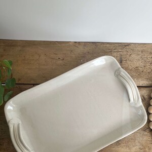 May include: A white ceramic serving tray with two handles on each side. The tray is on a wooden surface.