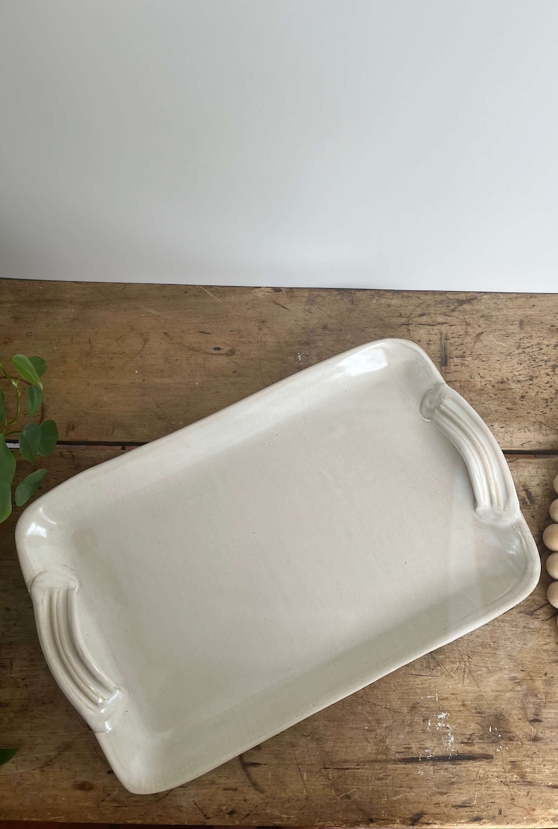 May include: A white ceramic serving tray with two handles on each side. The tray is on a wooden surface.