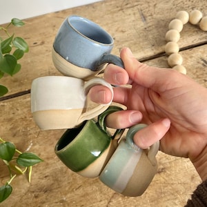 May include: Four small ceramic mugs in shades of blue, white, green, and a light green with a tan base. Each mug has a rounded handle and a contrasting base color. The mugs are stacked together, showcasing their handmade quality.