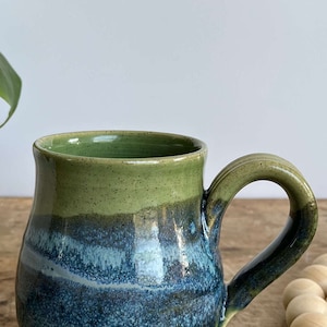 Op de afbeelding: Een groene en blauwe keramische mok met een rond lichaam en een handvat. De mok heeft een gespikkeld glazuur en een lichtblauwe streep.