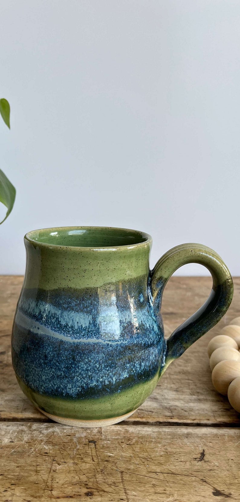 Op de afbeelding: Een groene en blauwe keramische mok met een rond lichaam en een handvat. De mok heeft een gespikkeld glazuur en een lichtblauwe streep.