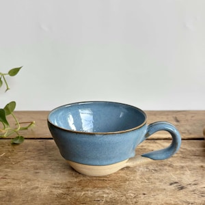 May include: A ceramic mug featuring a glossy blue glaze on the upper portion and a natural, unglazed beige base. The mug has a wide, rounded bowl shape with a thick, curved handle attached to the side, showcasing a distinct two-tone color design.
