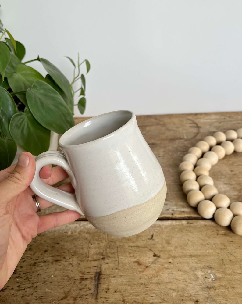 May include: A white ceramic mug with a rounded body and a handle. The mug has a light brown glaze on the bottom half.