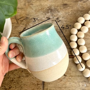 May include: A light blue, white, and beige ceramic mug with a handle. The mug is 4.5 inches tall and 3 inches wide.