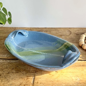 May include: An oval ceramic serving dish with a glossy blue glaze and a prominent diagonal band of mottled light green and white. The dish features two integrated, sculpted handles on opposite ends, creating a smooth, continuous form.