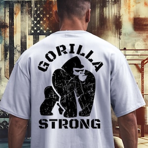 May include: A pale grey t-shirt featuring a black gorilla graphic and the text "GORILLA STRONG". The image is set in a gym, with weights and exercise equipment visible in the background.