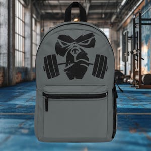 May include: Gray backpack featuring a black gorilla graphic holding a barbell. The backpack has a front zippered pocket, a top handle, and a mesh side pocket. The design is suitable for gym enthusiasts or anyone who appreciates fitness-themed accessories.