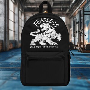 Op de afbeelding: Zwarte rugzak met een witte tijgerafbeelding en de tekst "FEARLESS" erboven en "ONLY THE STRONG SURVIVE" eronder. De rugzak heeft een voorvak en een handvat aan de bovenkant.