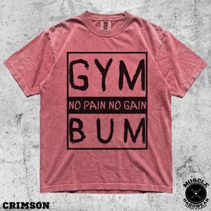 May include: A crimson short-sleeve t-shirt featuring a black rectangular graphic on the chest. The graphic contains the text GYM at the top, NO PAIN NO GAIN in the middle, and BUM at the bottom, all printed in a bold, distressed black font.