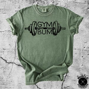 May include: A sage green short-sleeved t-shirt featuring a black graphic of a barbell with the text GYM BUM written in the center. The shirt has a crew neckline and a relaxed fit, with a visible brand label printed on the inside of the collar.