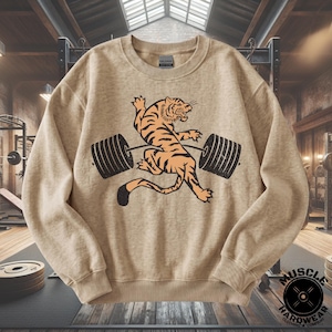Puede incluir: Una sudadera de cuello redondo color canela con un estampado gráfico de un tigre naranja y negro levantando una barra de pesas negra. La sudadera tiene mangas largas, cuello acanalado y puños acanalados. La etiqueta de la marca Gildan es visible.