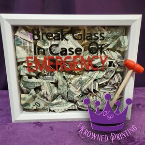 May include: A white framed box filled with crumpled dollar bills, with the text "Break Glass In Case Of Emergency" in black and red. A small wooden hammer with a red head rests on the side. A purple crown logo is in the bottom right corner.